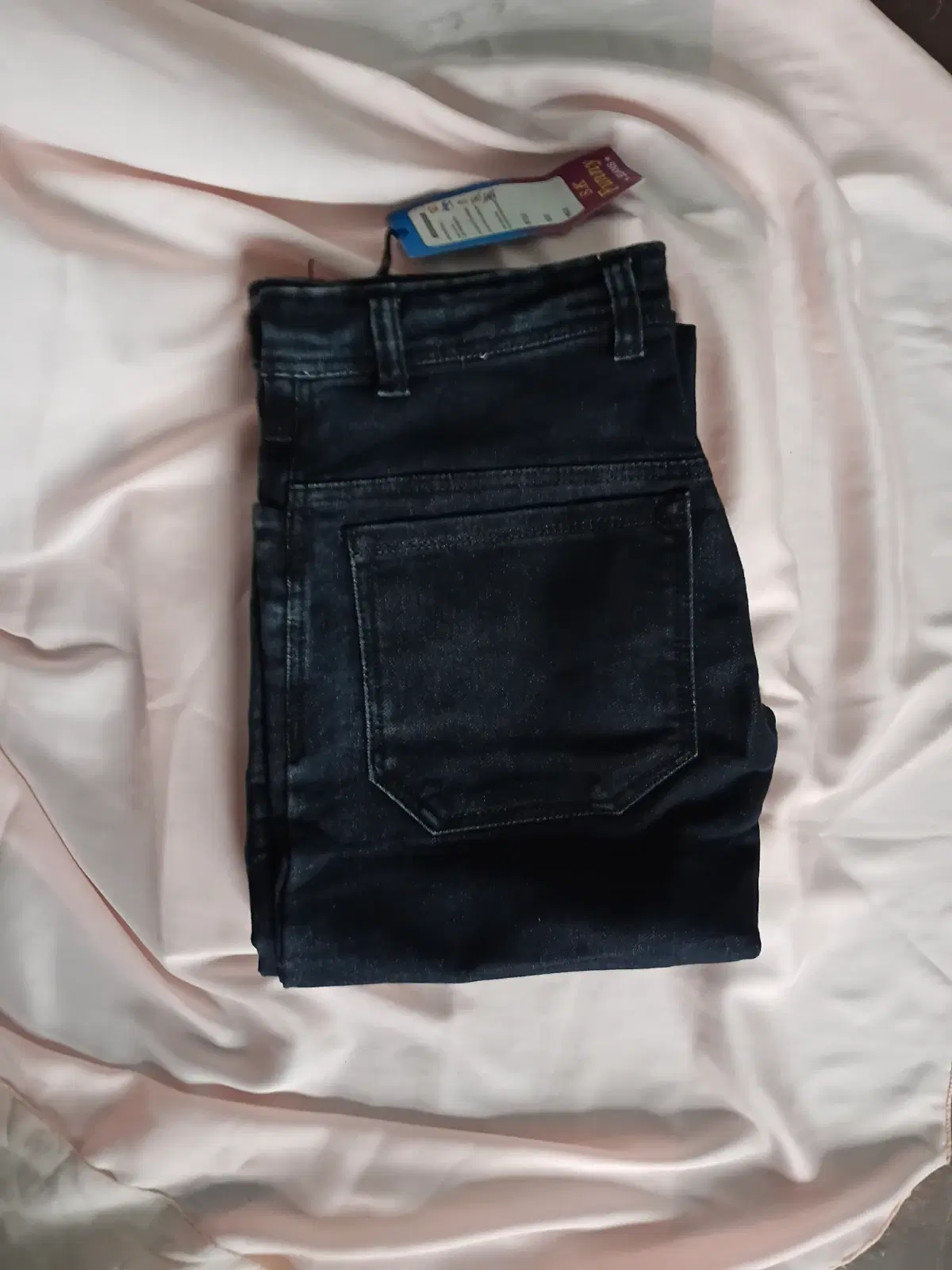 Folded dark wash denim jeans with back pocket detail and attached tag on a light fabric background