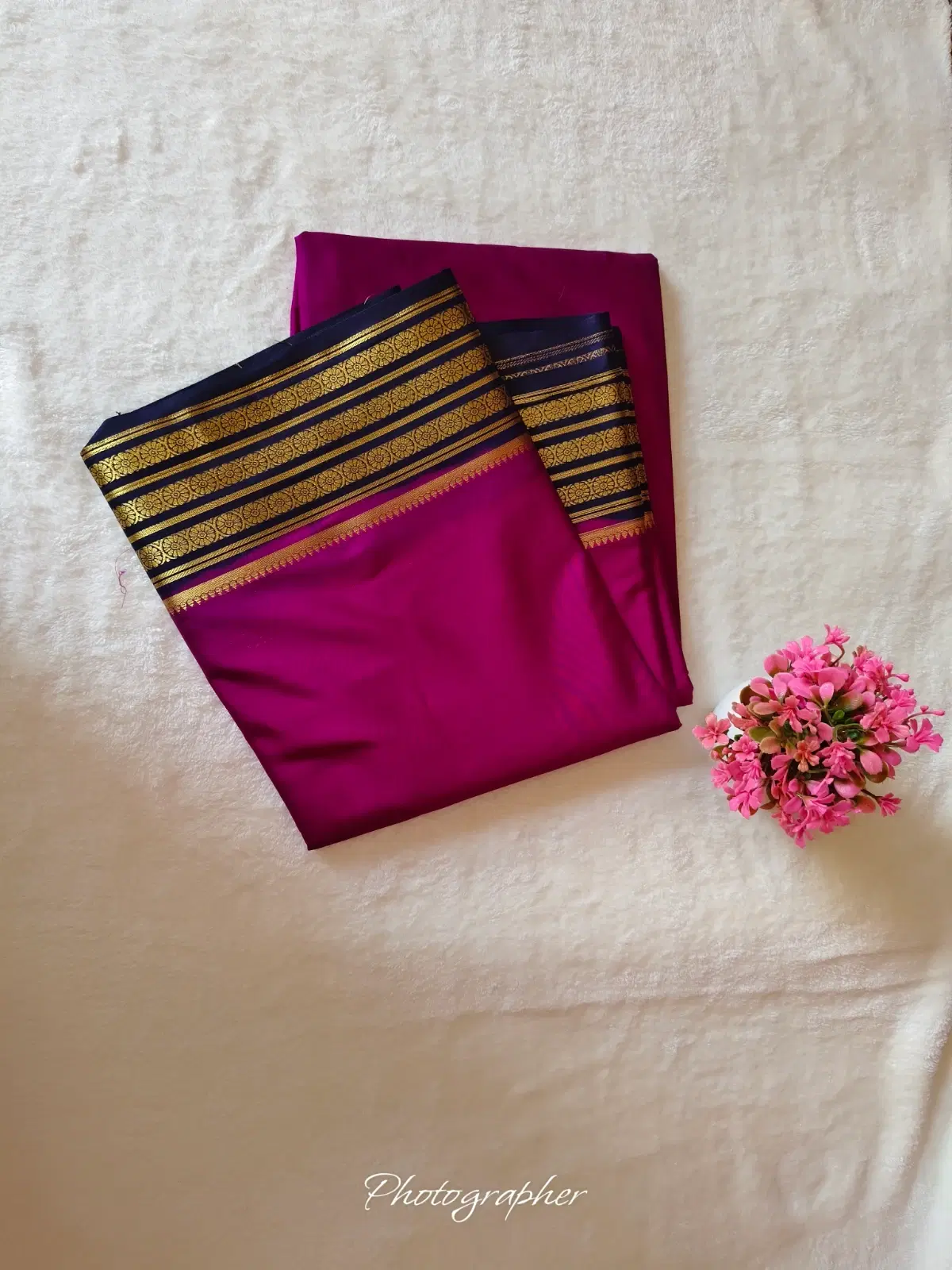 Folded magenta silk saree with black and gold zari border placed on a cream fabric beside pink flowers