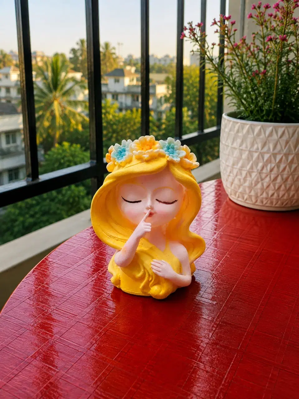 Yellow girl figurine with floral crown on a red table beside a white flower pot
