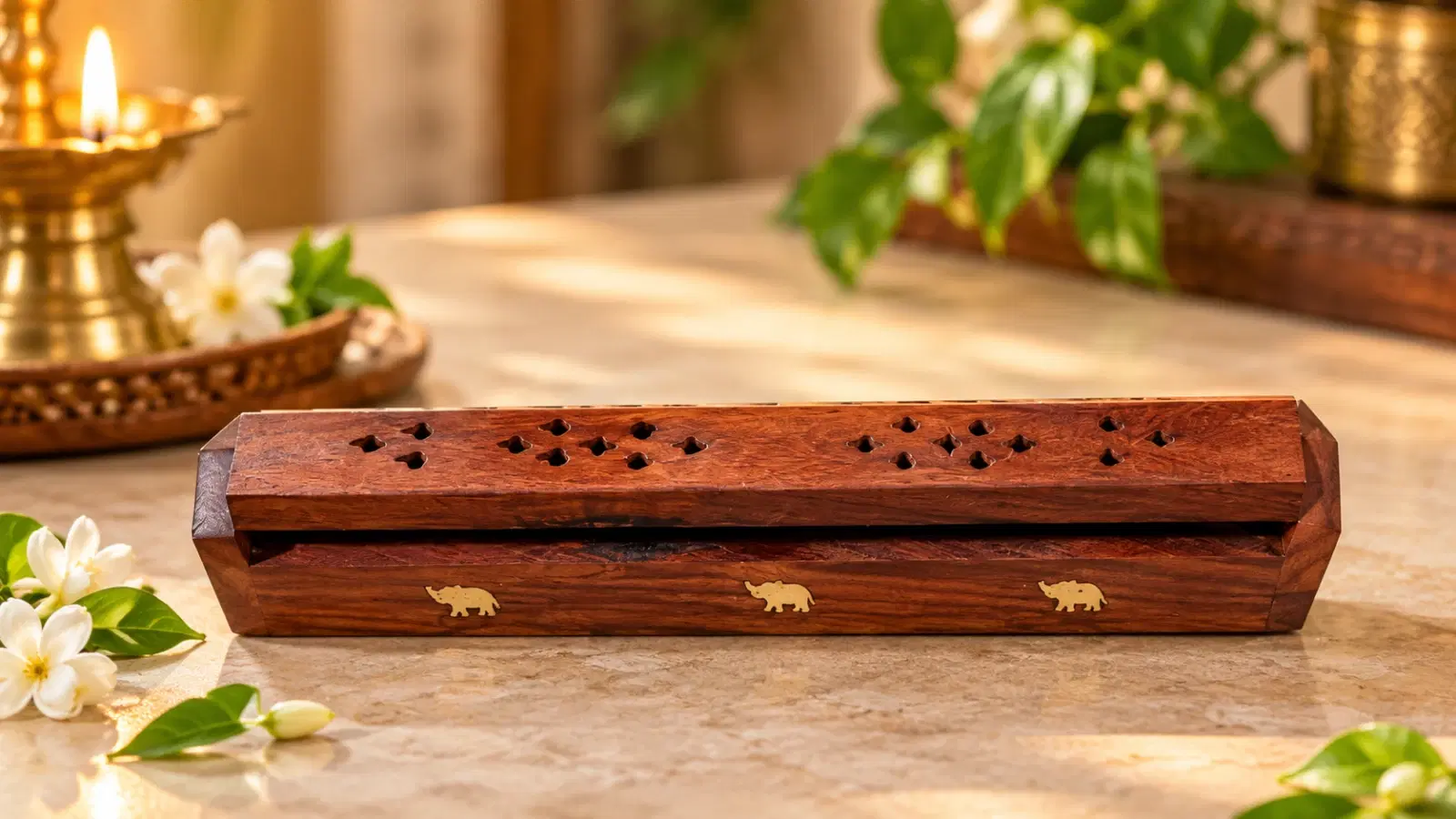 Wooden incense holder with carved top vents and elephant inlay, styled on a tabletop beside a lit brass diya