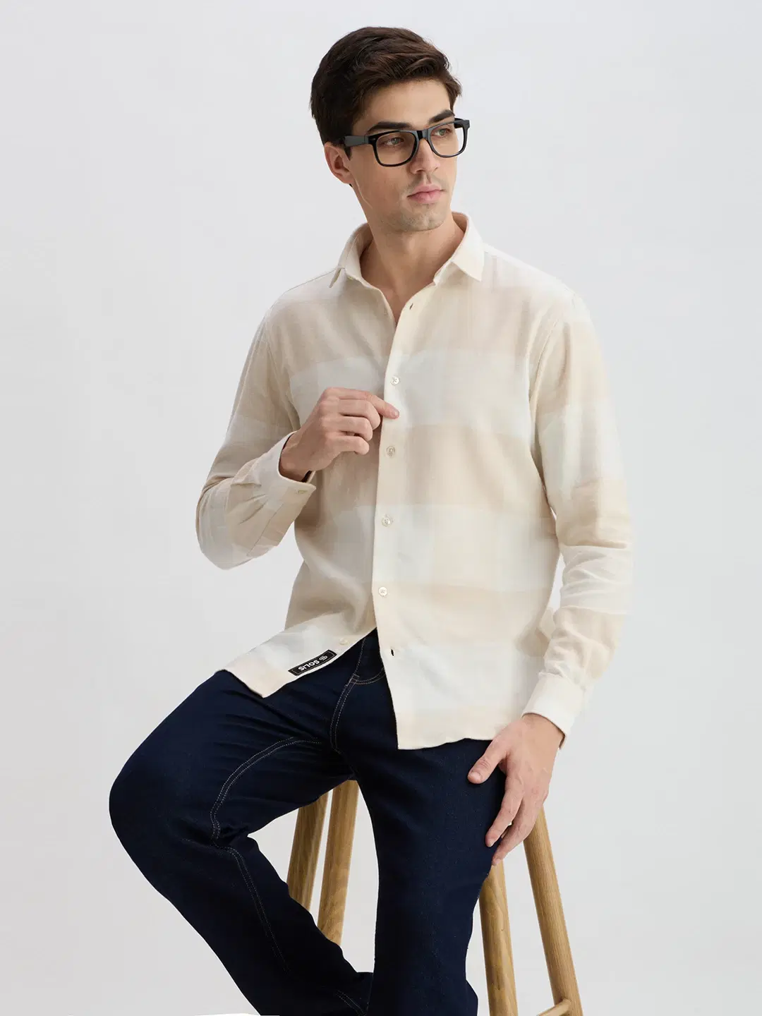Men's cream checked shirt styled seated on a wooden stool, angled side-front view