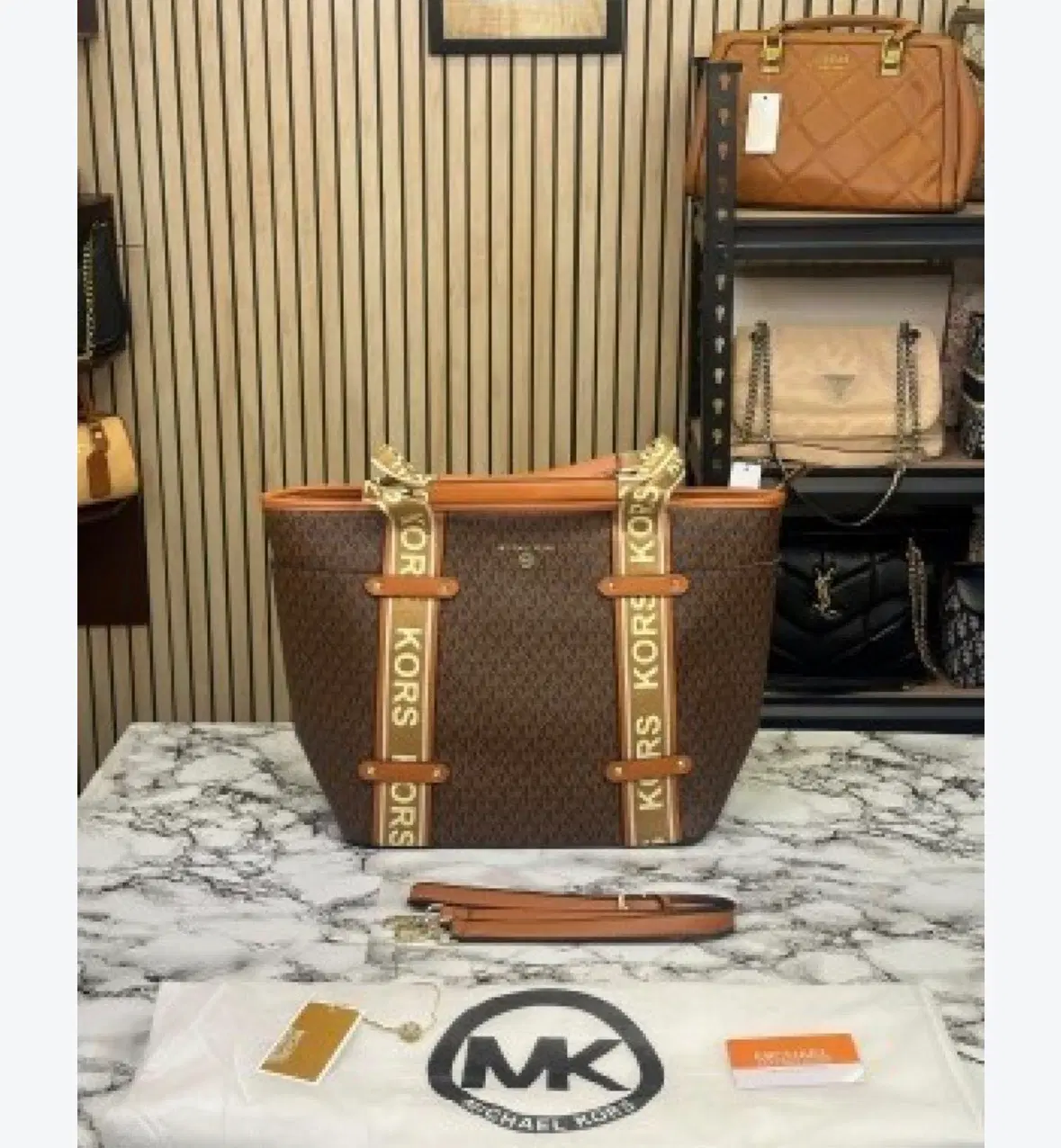 Brown MK monogram tote with tan trim and logo straps, shown front-facing with handles wrapped in protective cover