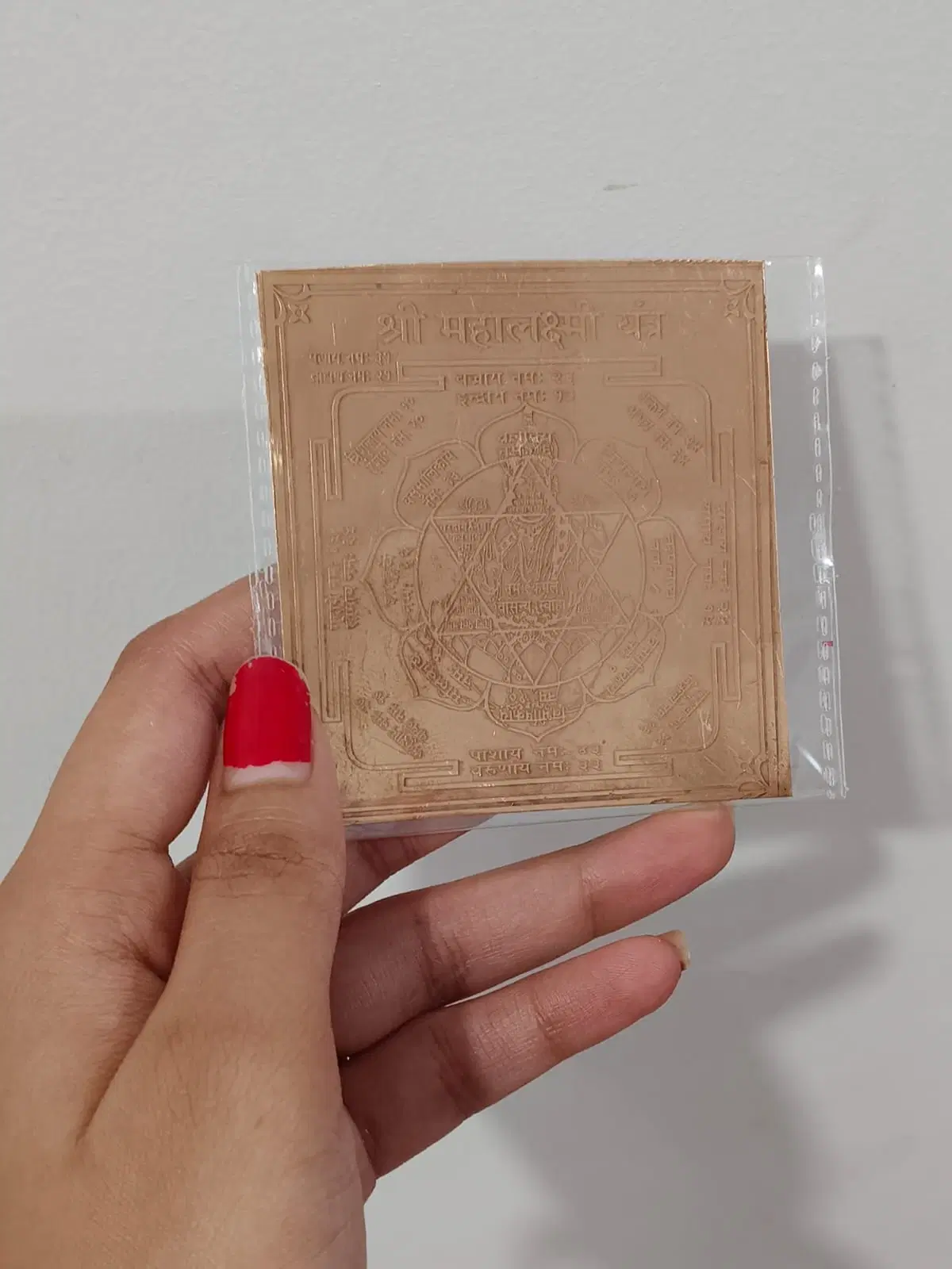 Hand holding a square copper-toned Shri Mahalakshmi Yantra plate sealed in a clear plastic sleeve